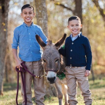 Two boys stand outdoors, smiling, with a donkey between them. One boy holds the donkey's reins. Trees are visible in the background.