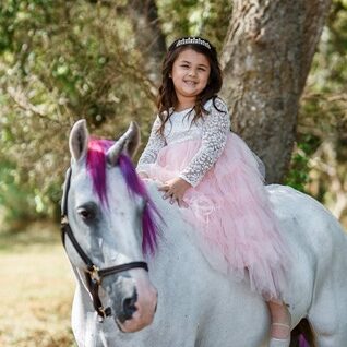 A young girl wearing a pink dress and a tiara sits on a white horse with purple accents in its mane, outdoors near a tree.